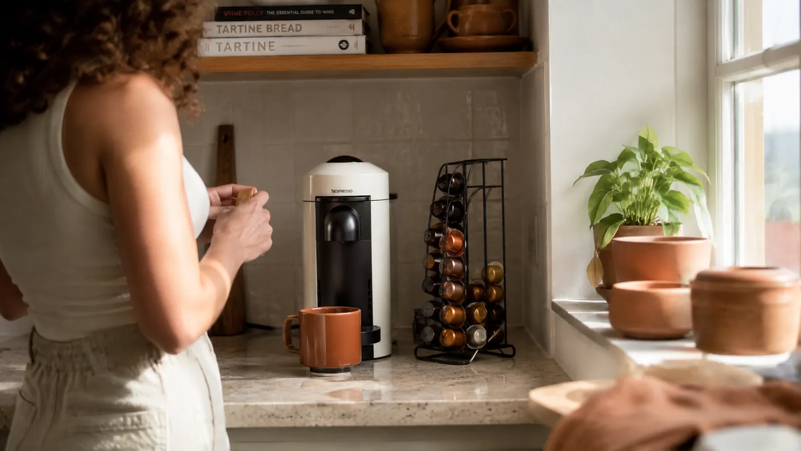 Compact coffee station in a small apartment kitchen with a narrow single-serve brewer, a small pod rack, and a white ceramic mug on a bone-colored tile counter